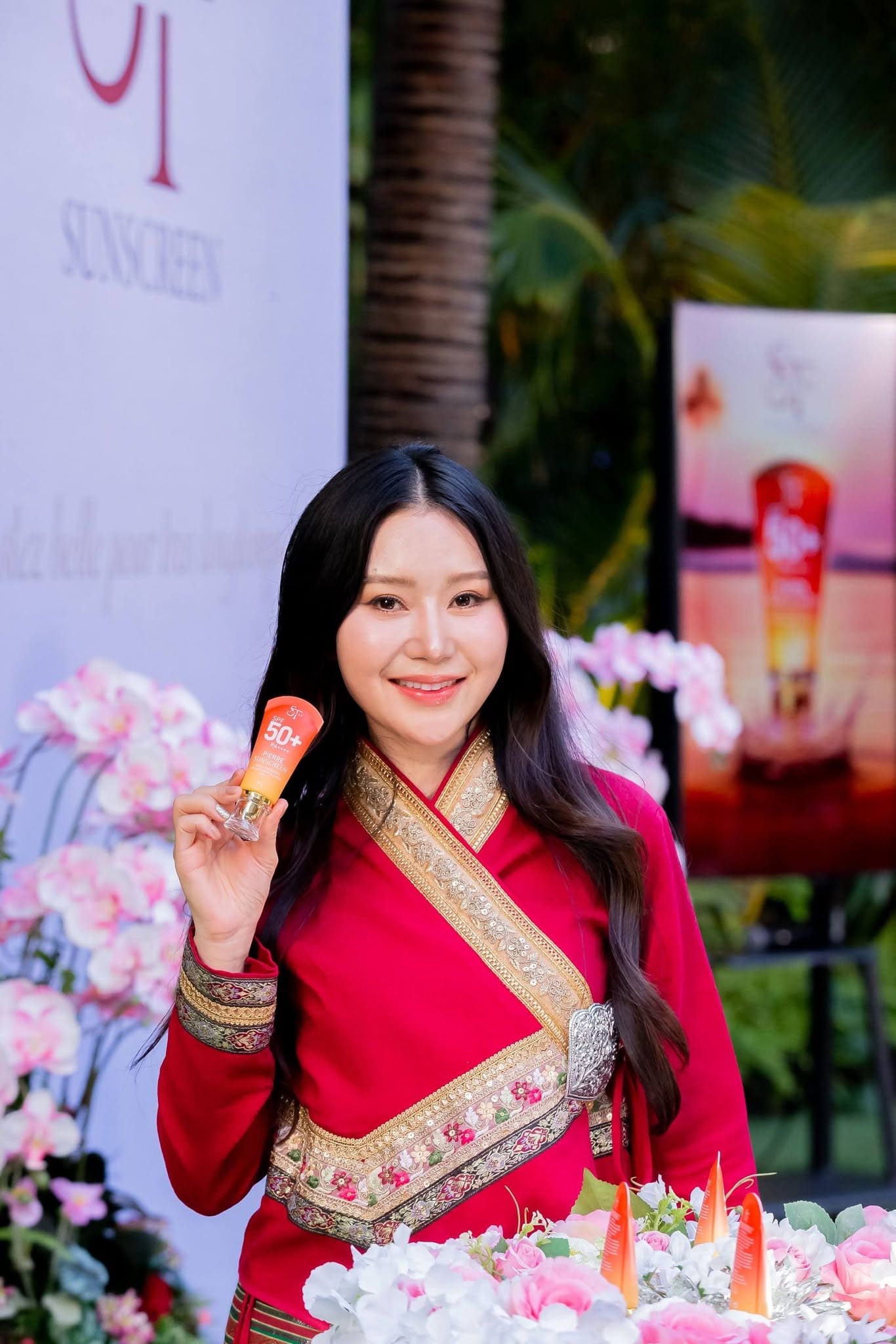 ภาพถ่ายครึ่งตัวหญิงสาวผมยาวสวมชุดไทยประยุกต์สีแดงตกแต่งแถบทอง ยิ้มพร้อมถือหลอดครีมกันแดด Pierre Sunscreen ท่ามกลางบรรยากาศงานดอกไม้