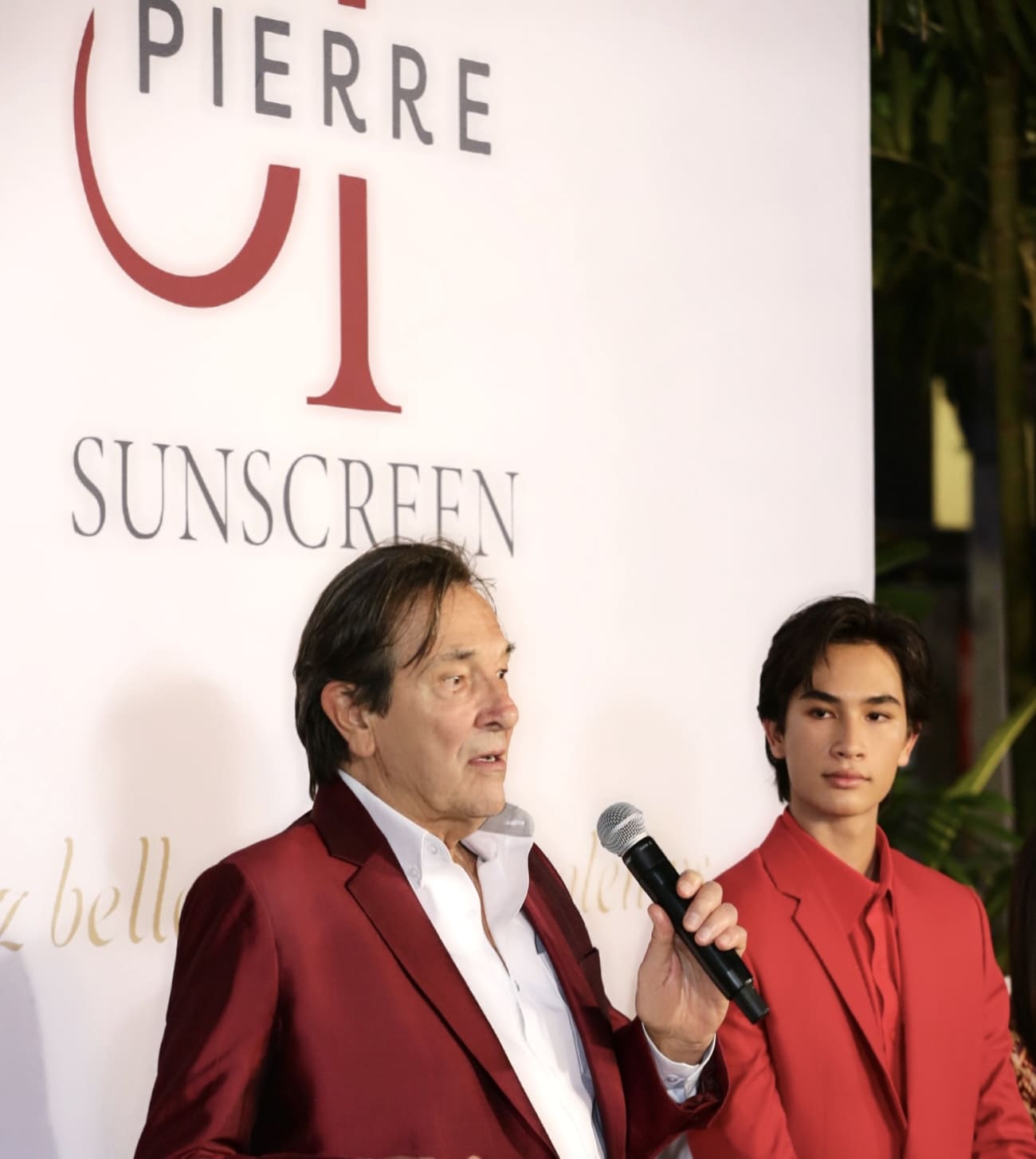 ภาพถ่ายผู้บริหารชายชาวต่างชาติในชุดสูทสีแดงเลือดหมูถือไมโครโฟนกำลังพูดบนเวที ยืนคู่กับพรีเซนเตอร์ชายสวมสูทสีแดงสด หน้าฉาก Backdrop โลโก้แบรนด์ Pierre Sunscreen