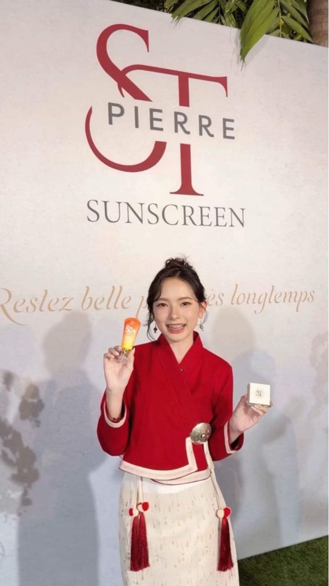 ภาพถ่ายเต็มตัวหญิงสาวสวมชุดไทยประยุกต์เสื้อสีแดง ยืนถือหลอดครีมกันแดด Pierre Sunscreen และกล่องบรรจุภัณฑ์หน้าฉาก Backdrop แบรนด์