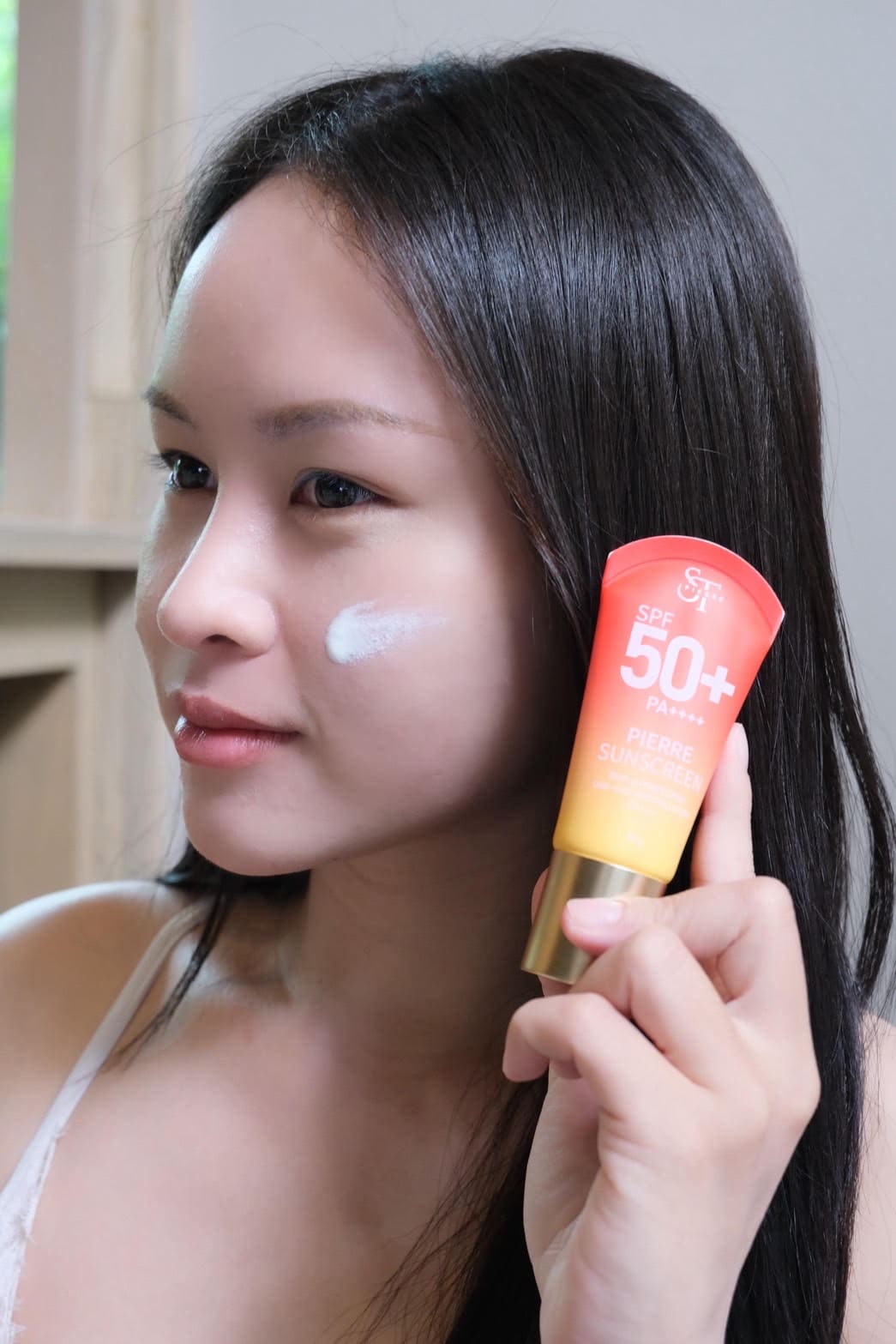 ภาพถ่ายระยะใกล้หญิงสาวถือหลอดครีมกันแดด Pierre Sunscreen พร้อมแต้มเนื้อครีมกันแดดสีขาวบนแก้ม แสดงการทดสอบเนื้อสัมผัสเบาบางบนผิวหน้า
