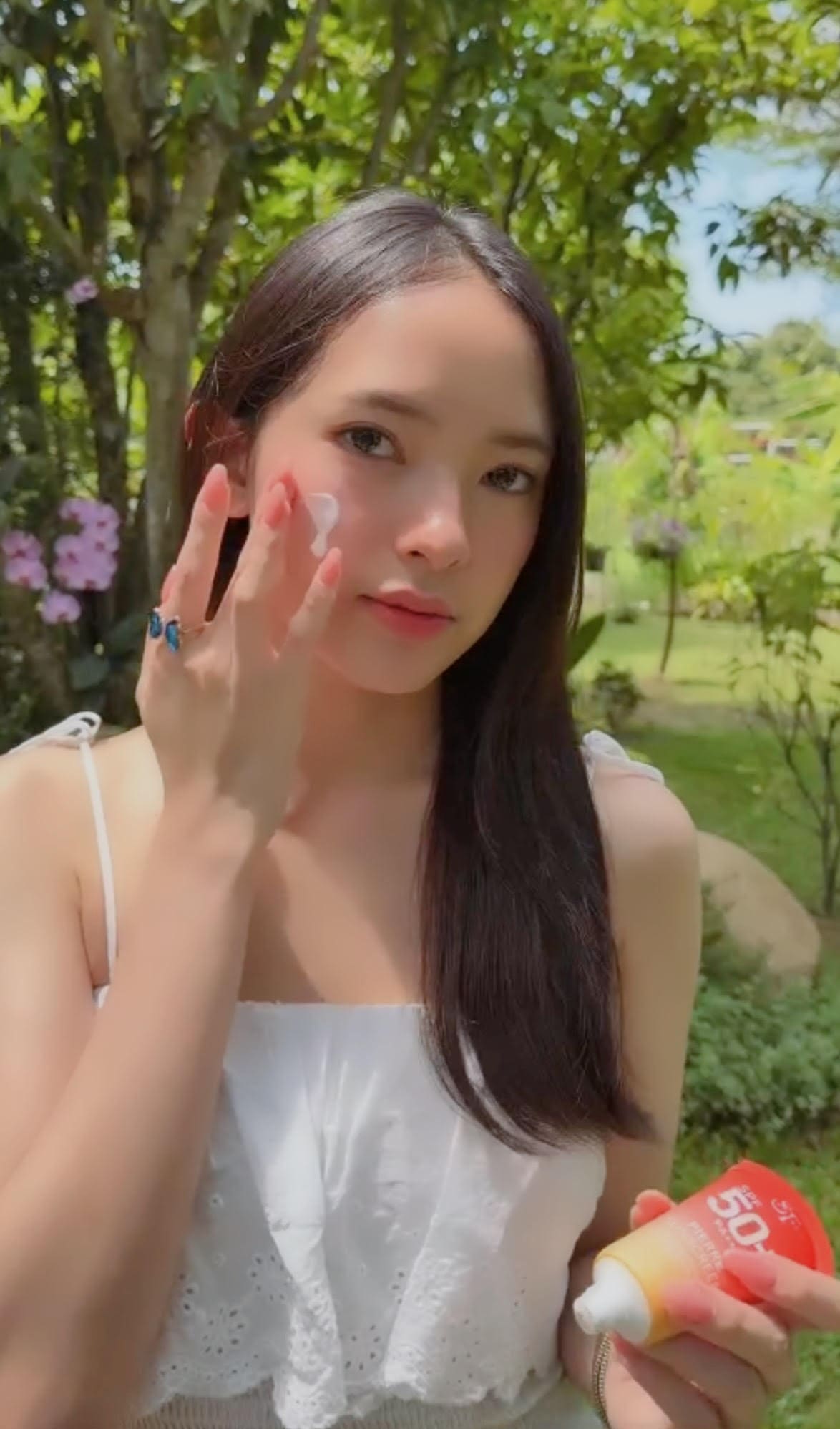 ภาพถ่ายหญิงสาวสวมเสื้อสีขาวกำลังใช้นิ้วเกลี่ยเนื้อครีมกันแดด Pierre Sunscreen บนใบหน้า มืออีกข้างถือหลอดสินค้า ท่ามกลางสวนธรรมชาติต่างจังหวัด