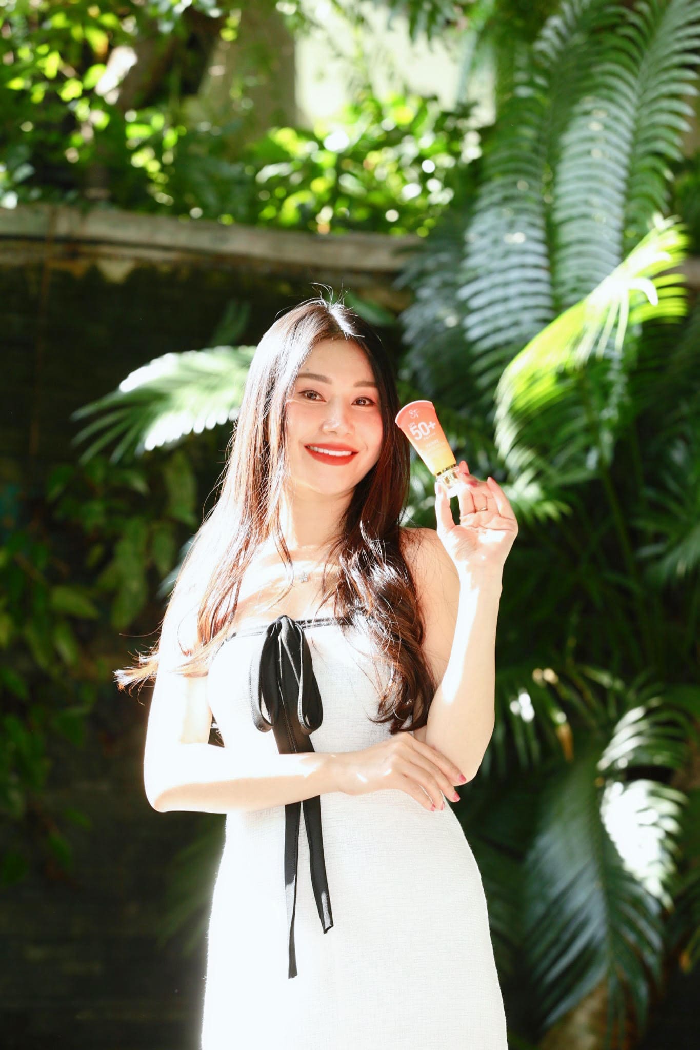 ภาพถ่ายครึ่งตัวหญิงสาวผมยาวสวมเดรสสีขาว ยิ้มสดใสพร้อมชูหลอดครีมกันแดด Pierre Sunscreen ในมือ ท่ามกลางแสงแดดและพื้นหลังต้นไม้สีเขียว