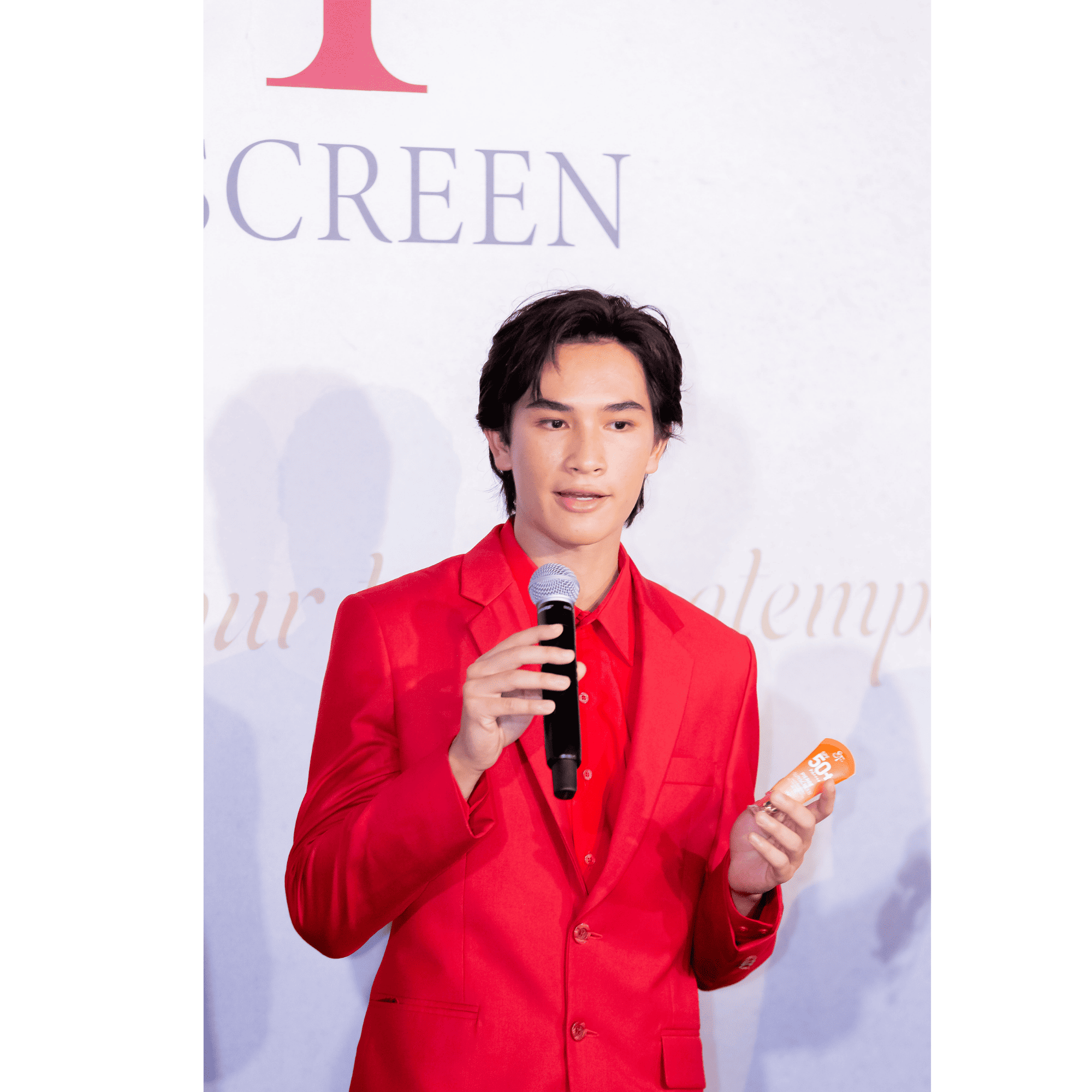 ภาพถ่ายครึ่งตัวพรีเซนเตอร์ชายสวมสูทสีแดง มือขวาถือไมโครโฟนพูดบนเวที มือซ้ายถือหลอดครีมกันแดด Pierre Sunscreen หน้าฉาก Backdrop งานเปิดตัวแบรนด์